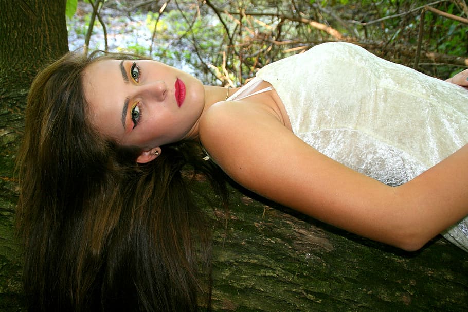 woman wearing white floral sleeveless dress lying on tree trunk during daytime