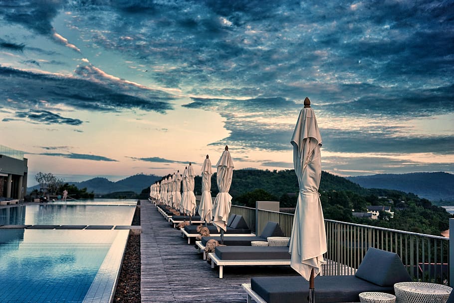 row of folded patio umbrella near pool, lounge chairs with patio umbrella beside swimming pool near body of mountains HD wallpaper
