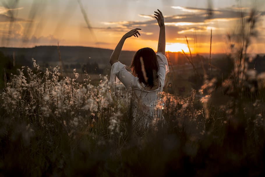 woman wearing white dress raising her two hands surrounded white petaled flowers during sunset, person in the middle of grass field during sunset