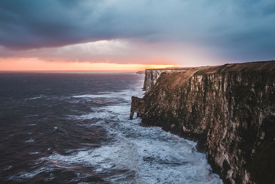 gray rocky mountain beside sea at golden hour, brown cliff near body of water under blue clouds with orange golden time horizon HD wallpaper