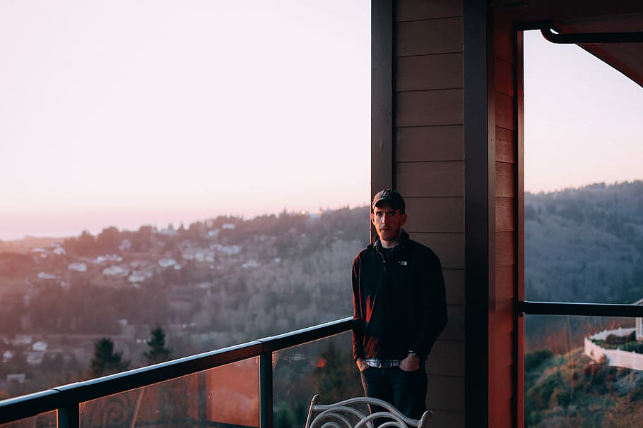 man standing near handrails during daytime, man wearing black jacket standing beside metal railing and behind brown louver wall overlooking plains at daytime HD wallpaper