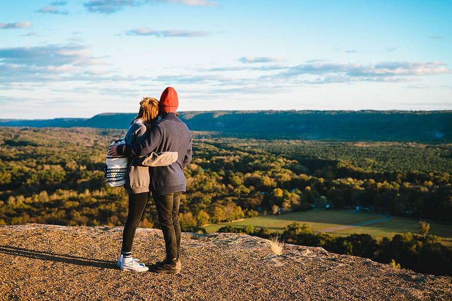 man and woman standing on mountain cliff in front of green trees, two person standing on mountain hugging each other HD wallpaper