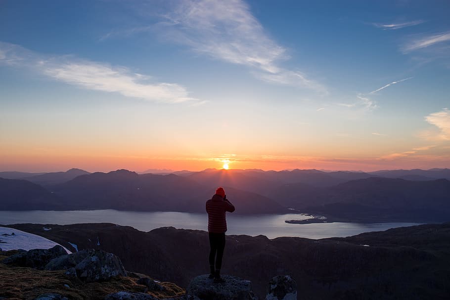 person standing on mountain ridge over lake under cloudy sky at sunset, silhouette photo of man standing on rock during sunset HD wallpaper