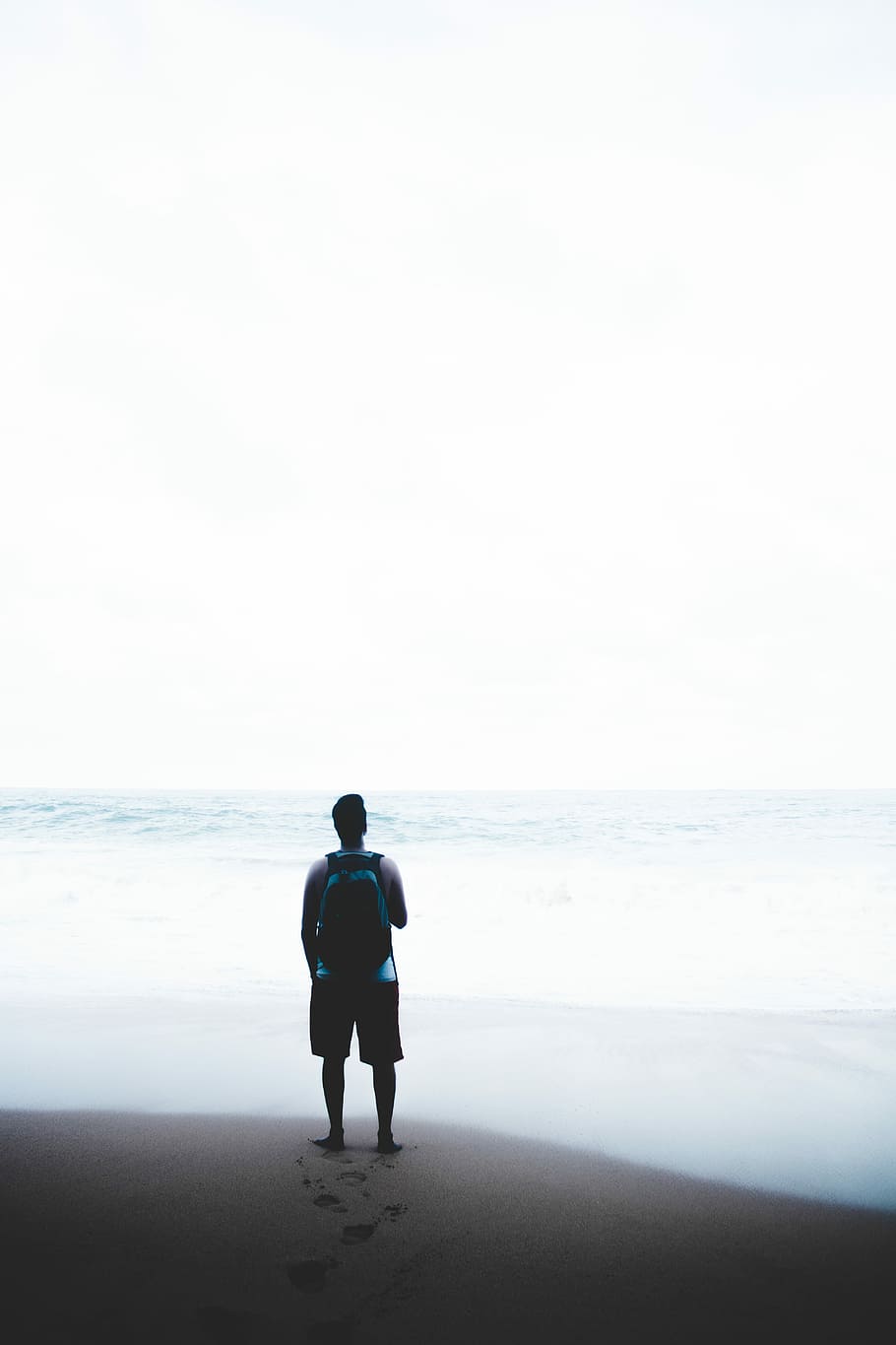 man standing in front of body of water while carrying blue and black backpack, man standing on brown shore near body of water under white sky HD wallpaper