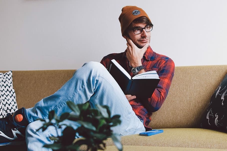 man sitting on sofa while holding book, man sitting on sofa holding book and his chin beside wall HD wallpaper