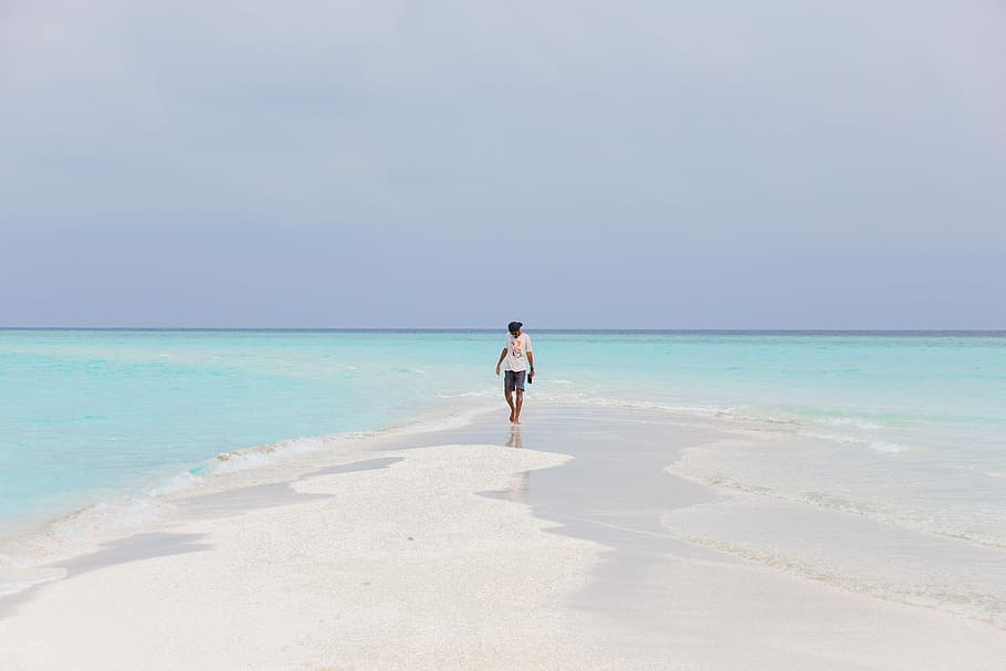 person wearing white shirt walking on seashore near blue body of water under blue sky during daytime, man in white crew-neck shirt walking on seashore during daytime HD wallpaper