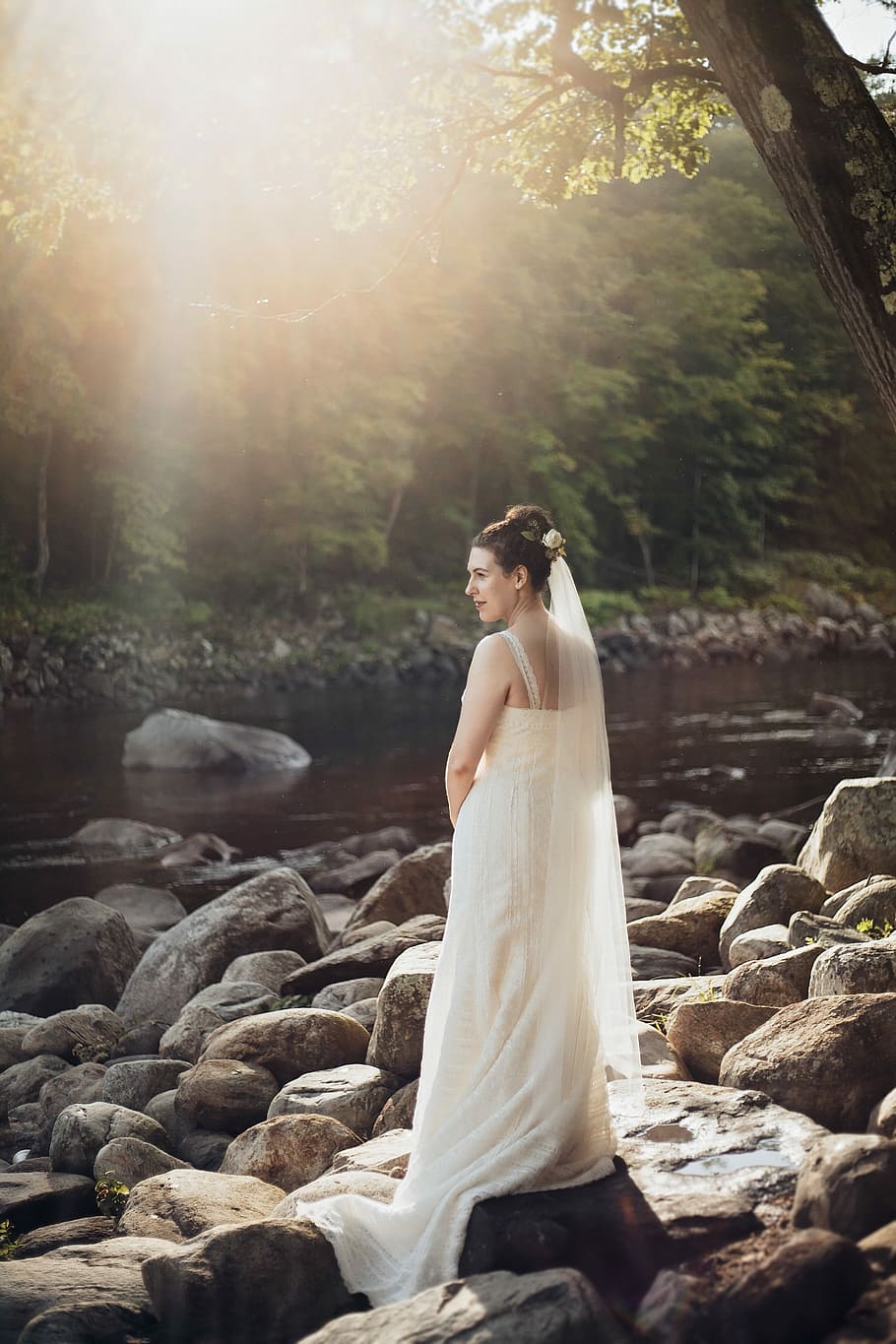 woman standing on gray rock near lake during sunset, woman wearing white sleeveless wedding gown standing on rocks near body of water