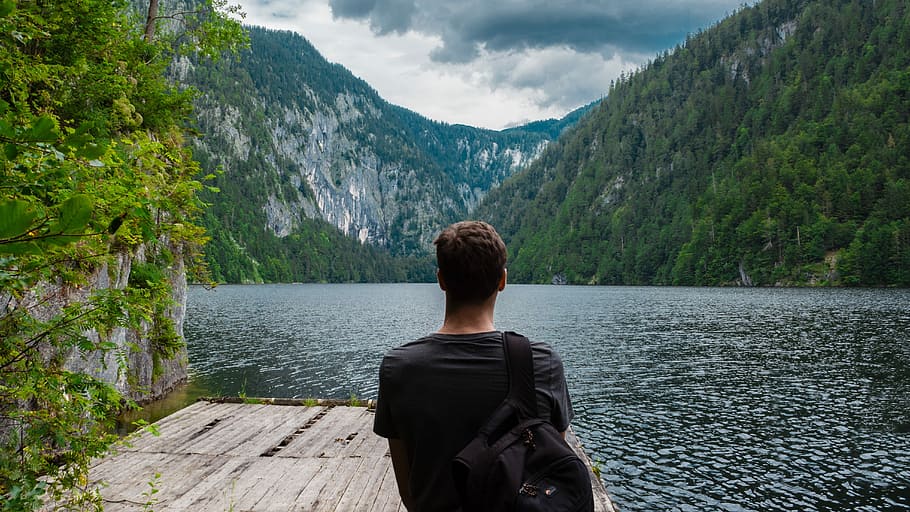 man in black shirt standing on dock beside lake surrounded by mountains HD wallpaper