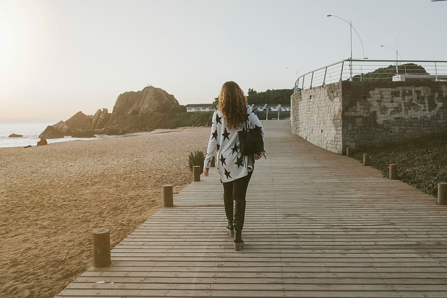 woman in white and black star print long-sleeved shirt and black denim jeans walking on brown wooden pathway near body seashore, HD wallpaper