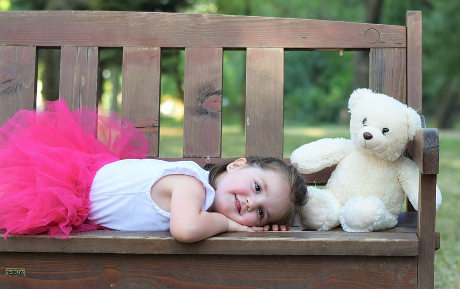 girl lying on brown bench beside bear plush toy at daytime, kid