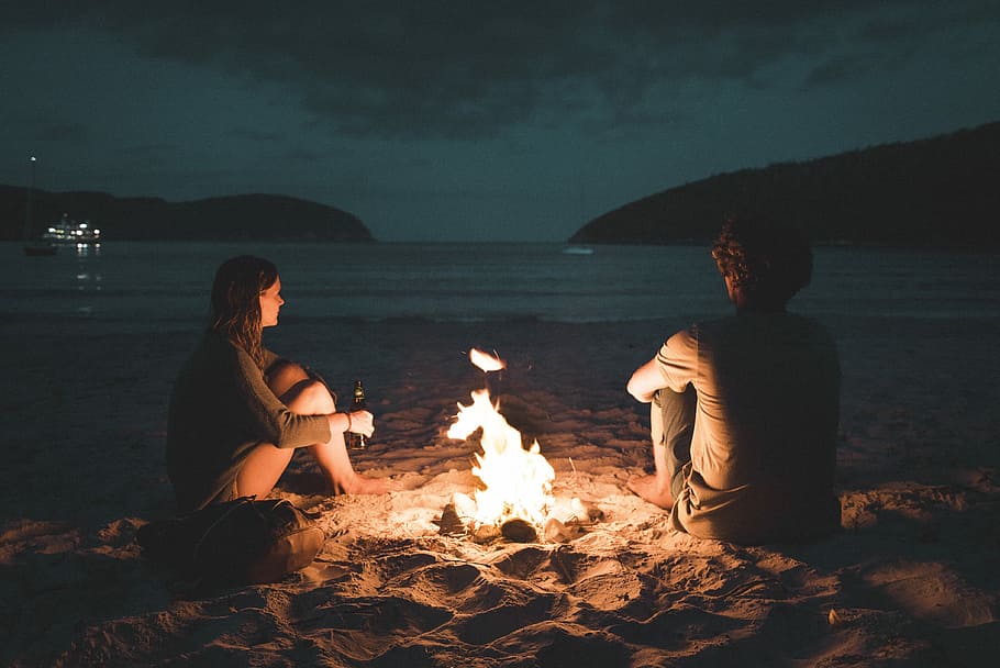 man and woman with bone fire sitting on seashore, bonfire between man and woman near sea during nighttime