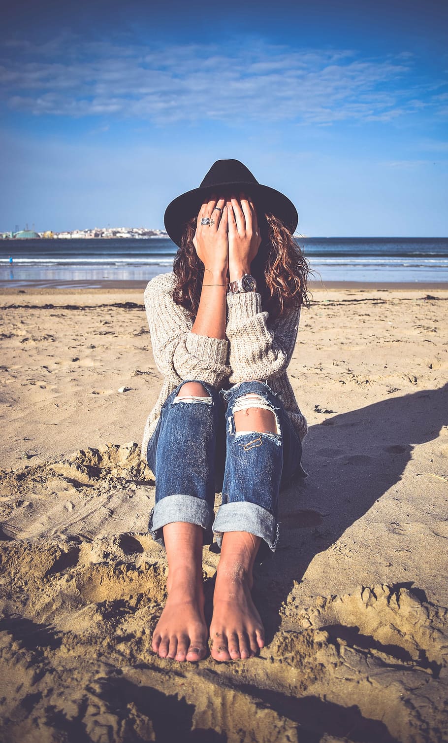 woman in gray sweater and blue denim distressed pants covering face with hands sitting on sand near body of water, HD wallpaper