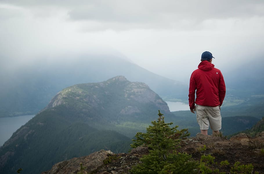 person wearing fitted cap and red jacket standing on peak across hills and body of water HD wallpaper