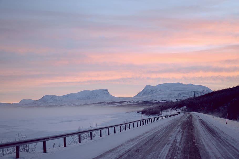 concrete road covered with snow taken at golden hour, snow-covered road near ocean and mountains HD wallpaper