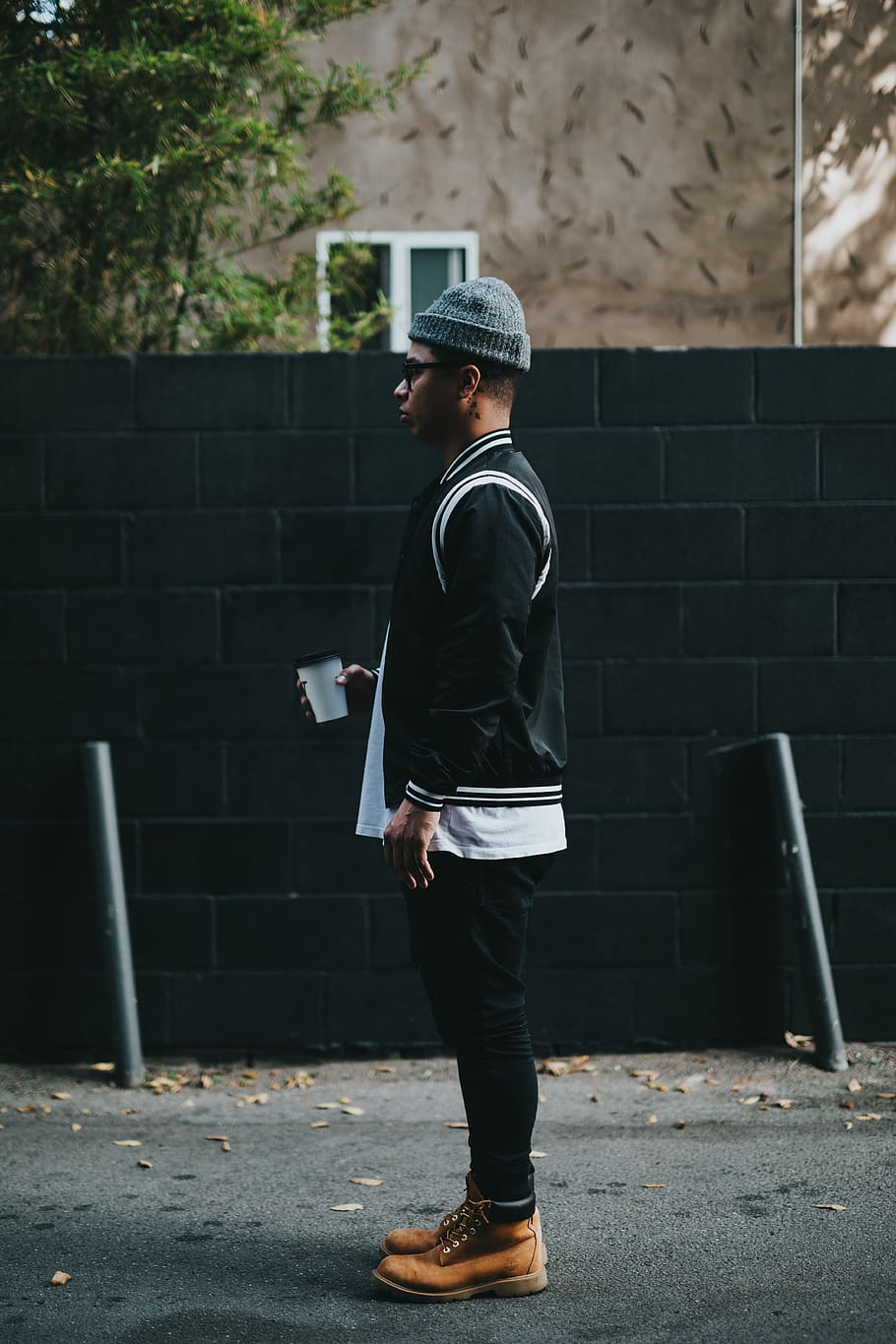 man standing near wall holding a cup looking forward, man wearing black and white jacket and black pants standing near black wall at daytime, HD wallpaper