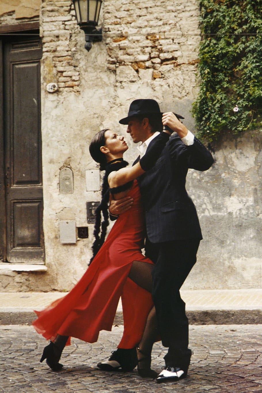 man in black suit and woman in red slit dress dancing beside grey concrete wall