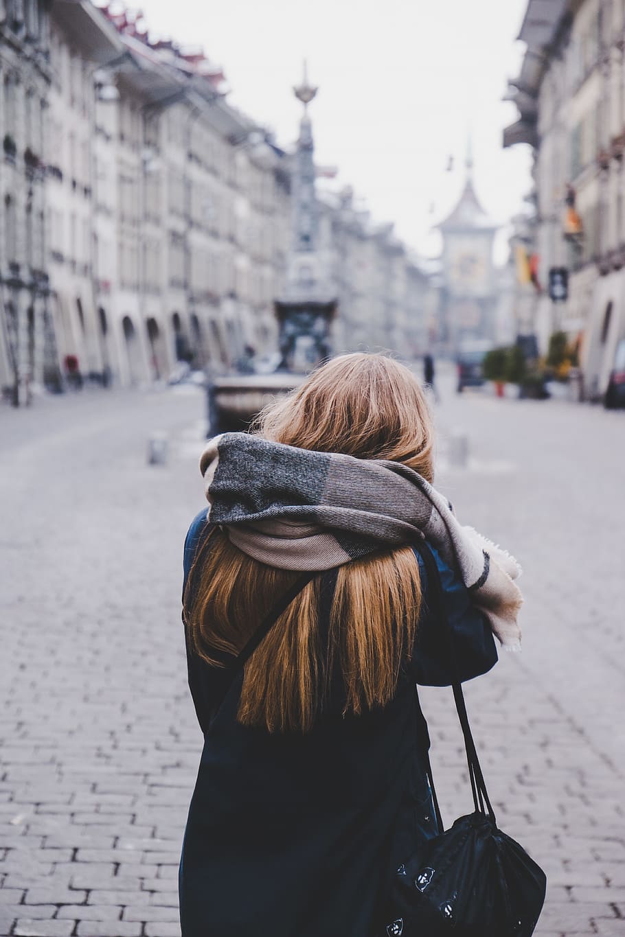 woman standing on sidewalk during daytime, woman wearing scarf standing in open area surrounded by buildings