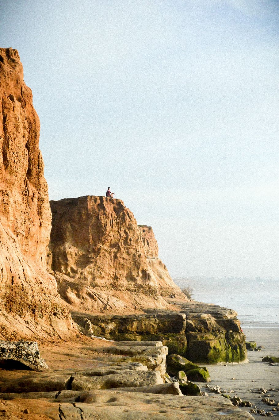 man on cliff near water, person sitting on brown cliff, rocks HD wallpaper