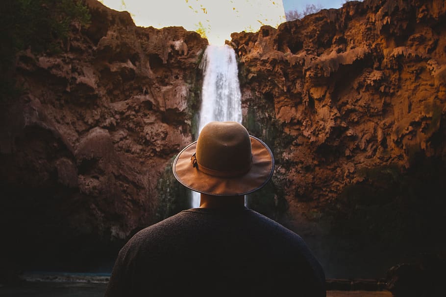 person facing waterfalls, man wearing brown hat standing fronting waterfalls HD wallpaper