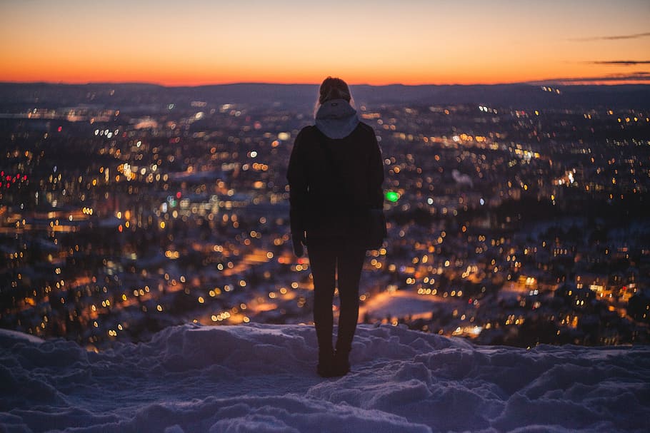 person standing on mountain looking on lights, woman wearing hoodie standing near the edge of snow covered cliff facing the city during night HD wallpaper