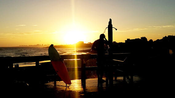 silhouette of man on terrace, silhouette of man standing on the shower near surfboard and sea shore during sunset HD wallpaper