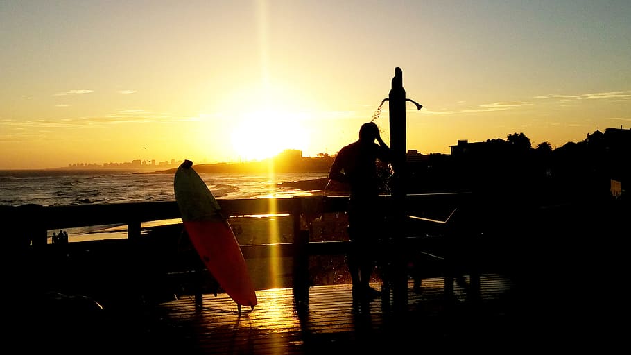 silhouette of man on terrace, silhouette of man standing on the shower near surfboard and sea shore during sunset HD wallpaper