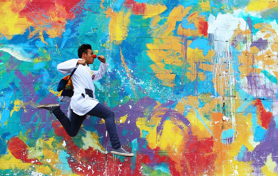 A dark-skinned male leaping along the sidewalk in front of an abstractly painted wall., man beside assorted-color wall mural during daytime HD wallpaper