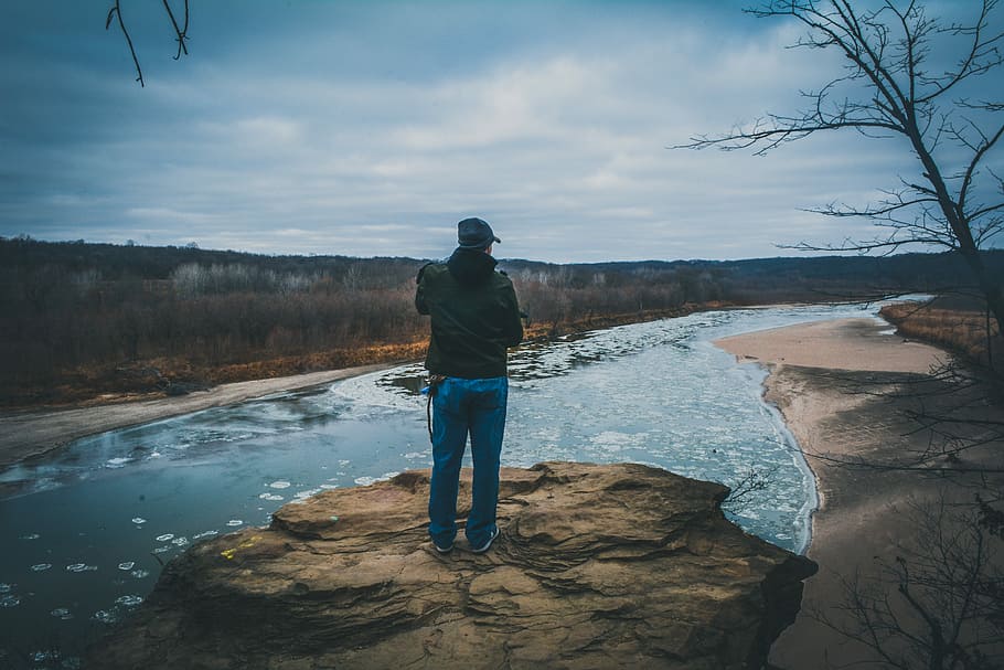man standing on mountain top, man in blue denim pants standing on brown rock looking at the river HD wallpaper
