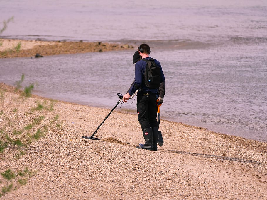 man using metal detector and holding shovel walking on beach