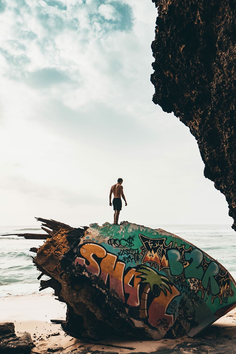 man standing on rock near beachline, man wearing black shorts standing on big rock near body of water under white sky during daytime, HD wallpaper