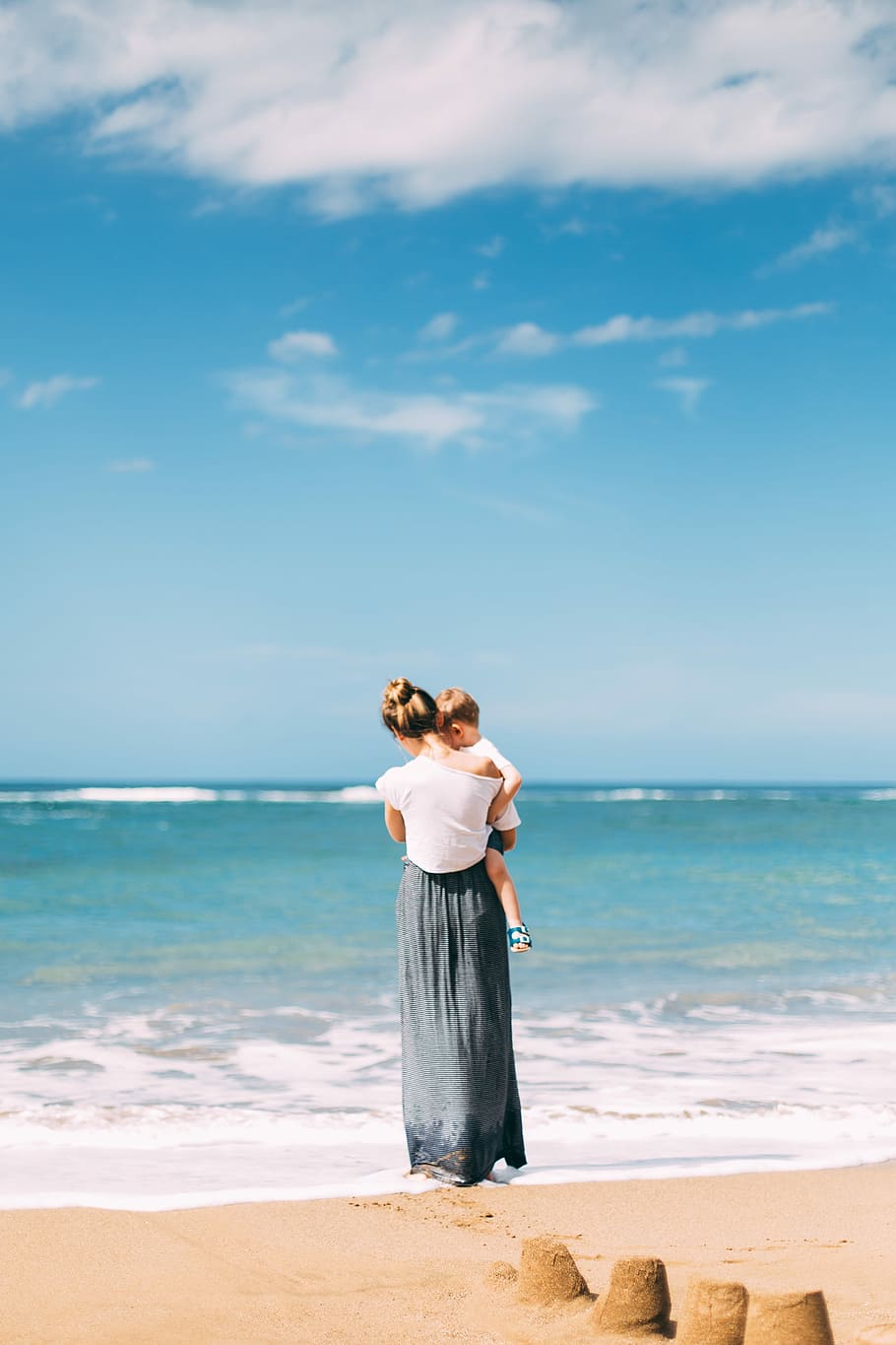 woman carrying toddler on seashore, woman carrying baby while standing on seashore during daytime