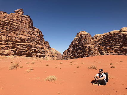 Man Wearing White Shirt and Black Pants Sitting on Soil Behind Rock Formation Mountain HD wallpaper