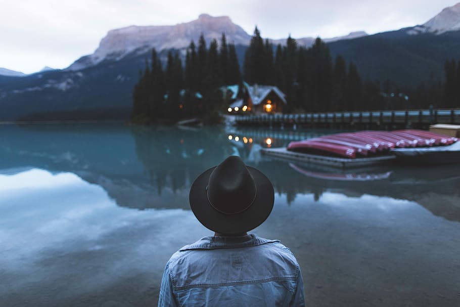 man standing infront of water with boats on dock with house nearby, person looking on green mountain