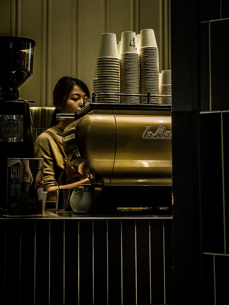 woman wearing gray sweatshirt standing near brown espresso machine with cups, woman operating gray metal espresso maker inside well lighted room HD wallpaper