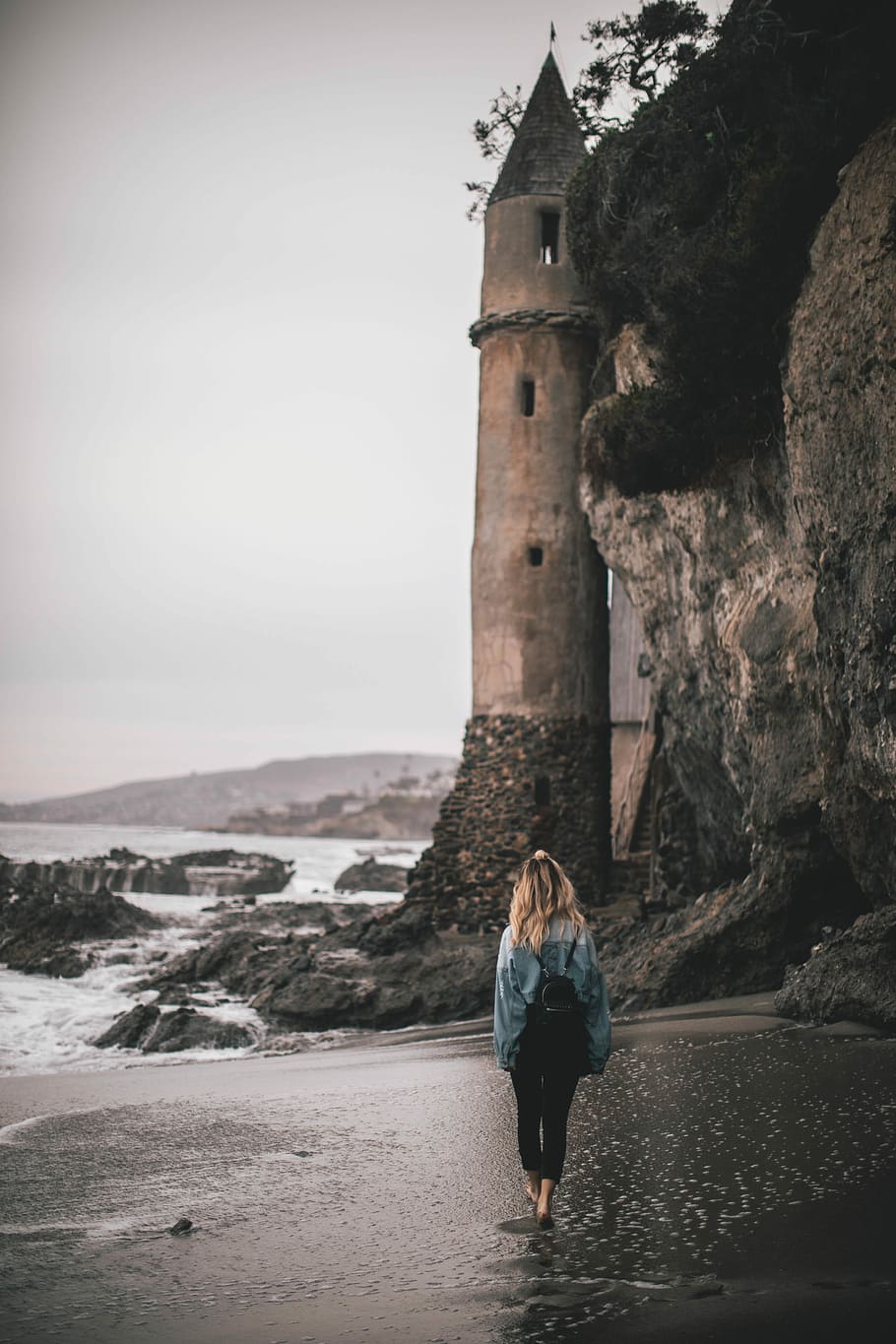 woman with backpack standing by the seashore near brown lighthouse, woman walking in between seashore and hill towards gray concrete tower HD wallpaper