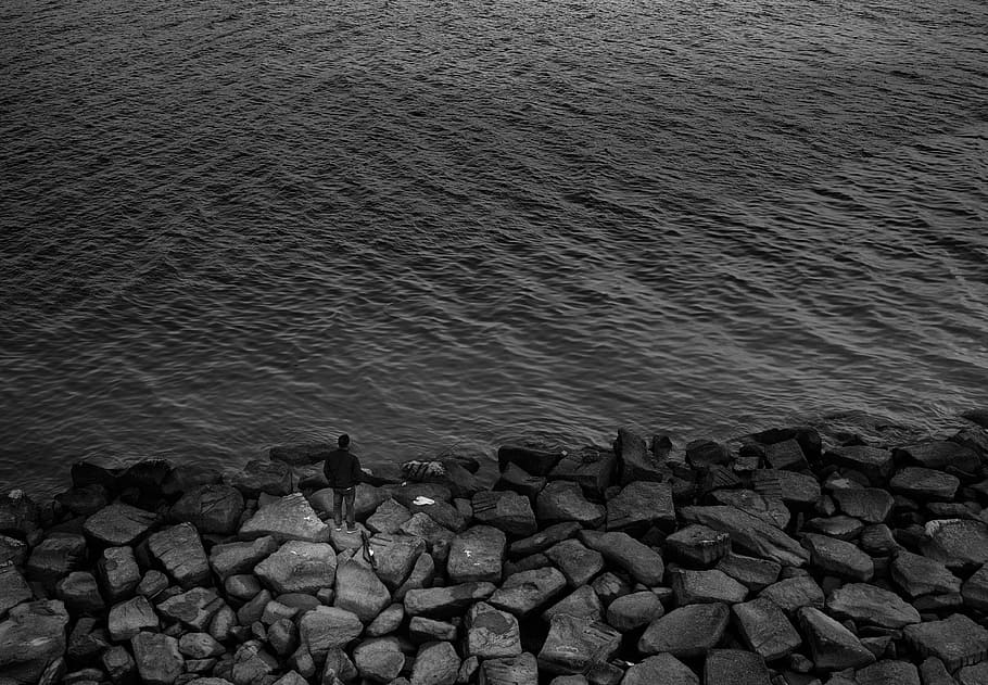 grayscale photography of man standing on rocks near body of water