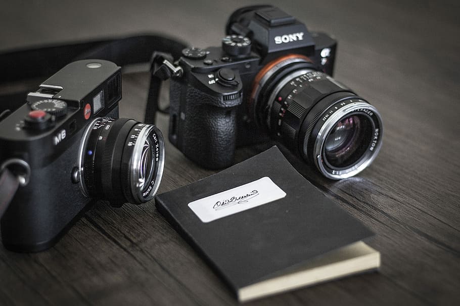 black Sony mirrorless camera near other mirrorless camera and notebook, selective focus photography of two black Sony DSLR cameras both on brown wooden surface HD wallpaper