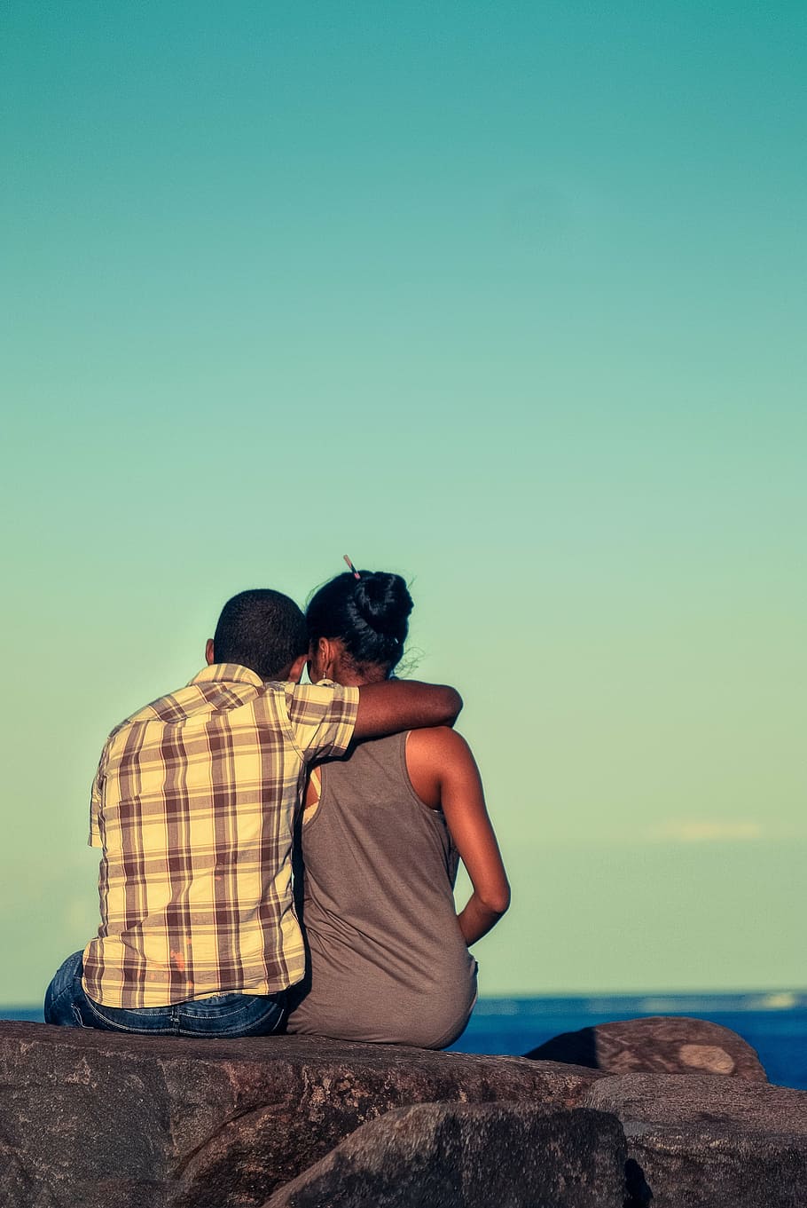 man wearing beige and gray plaid shirt hugging woman in gray tank top sitting on gray concrete ledge HD wallpaper