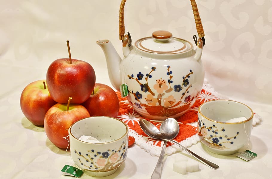 four red apples near white and multicolored ceramic teapot near two white ceramic teacups and two spoons HD wallpaper