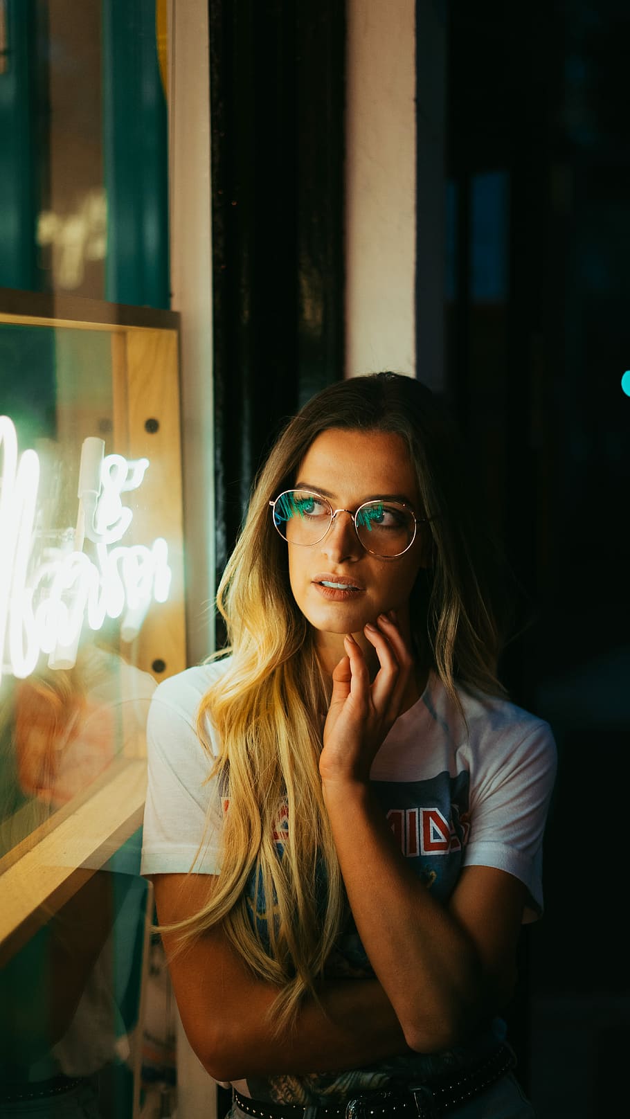 woman leaning on white wall with eyeglasses taking photo inside dark room, tilt-shift photography of woman standing beside LED light signage, HD wallpaper