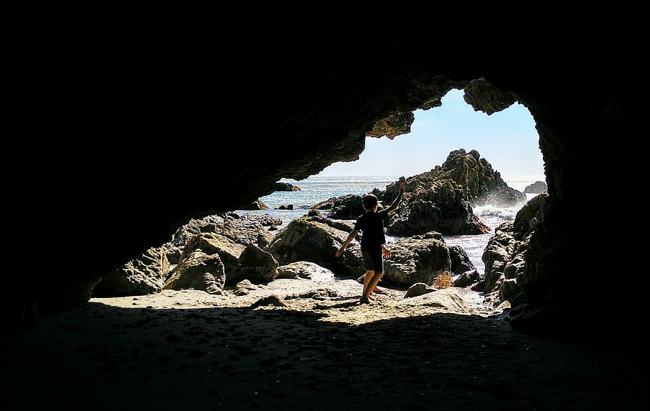 boy standing near stone, person standing in front of cave near body of water HD wallpaper