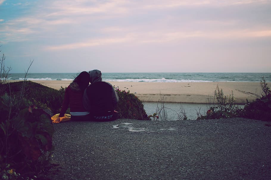 man and woman sits on gray asphalt way over ocean under cloudy sky during daytime, couple sitting in front of beach during daytime HD wallpaper