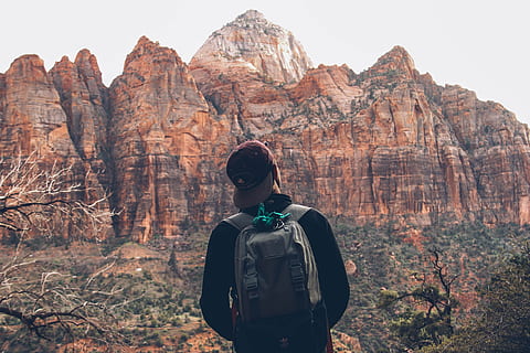 man looking at brown stone mountain, person wearing grey backpack standing near mountains HD wallpaper