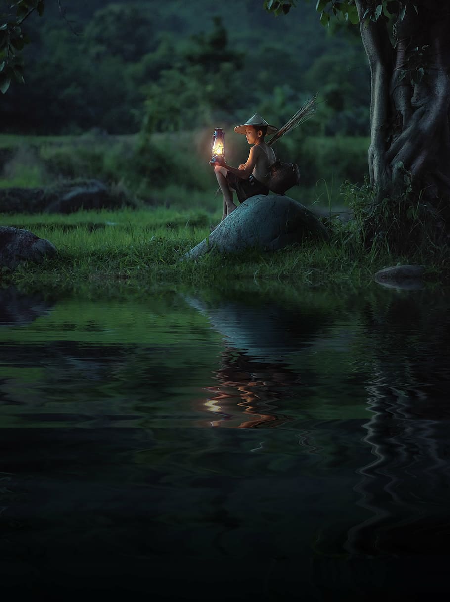 boy holding kerosene lamp sitting on stone near river, background