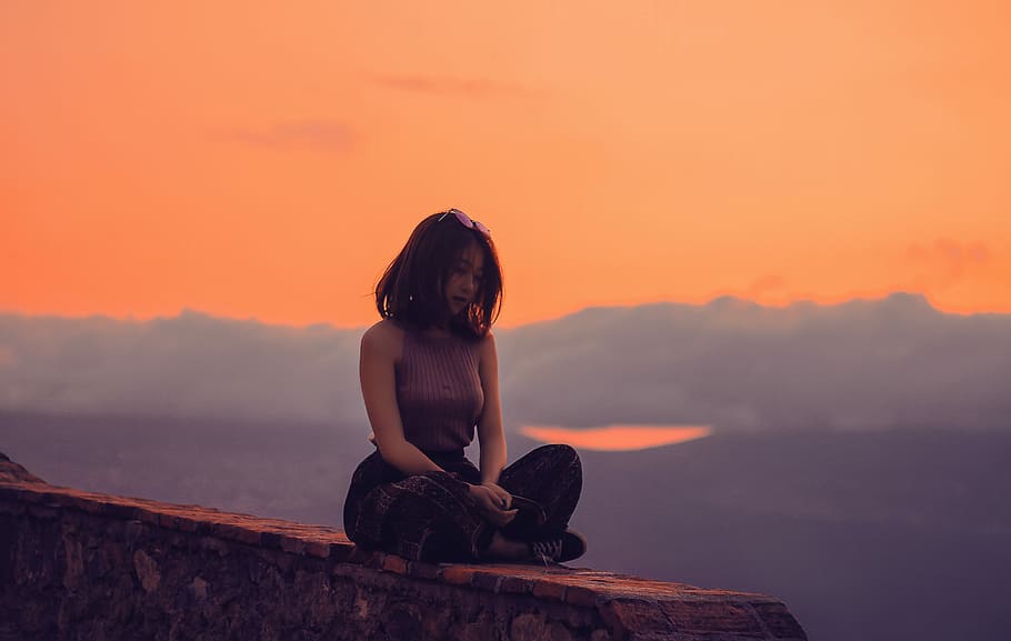 woman sitting on a cliff, woman in purple sleeveless top and black pants sitting on the edge of cliff at sunset HD wallpaper