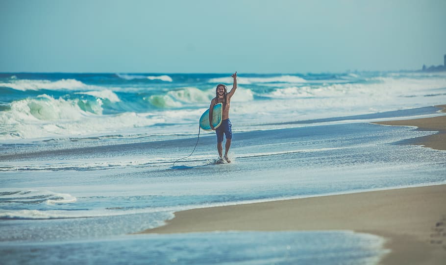 man carrying surfboard at beach, person waving and holding white and blue surfboard HD wallpaper