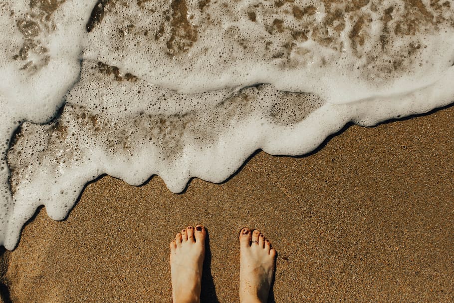 person's feet on seashore, person standing on beach sand near ocean waves