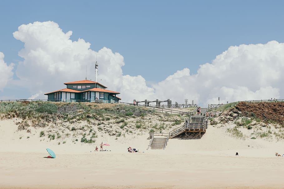 gray house under white clouds, people in beach near brown stairs in distant of blue and brown structures HD wallpaper