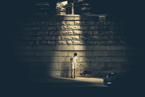 man in white shirt and white bottoms standing beside gray concrete wall while holding dog leash near car during nighttime HD wallpaper