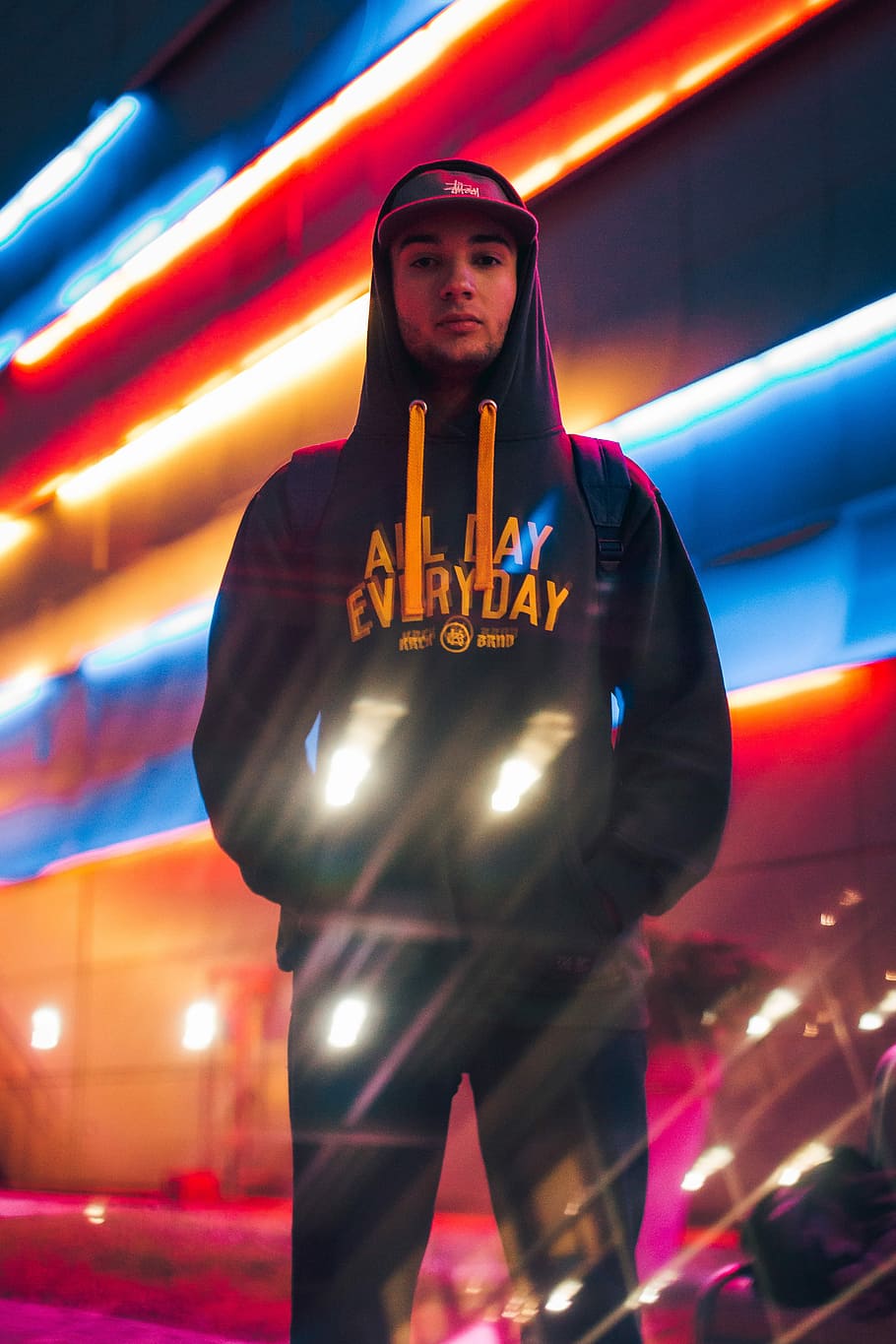 man standing behind building with red and blue neon lights, man standing wearing hoodie near building with lights HD wallpaper